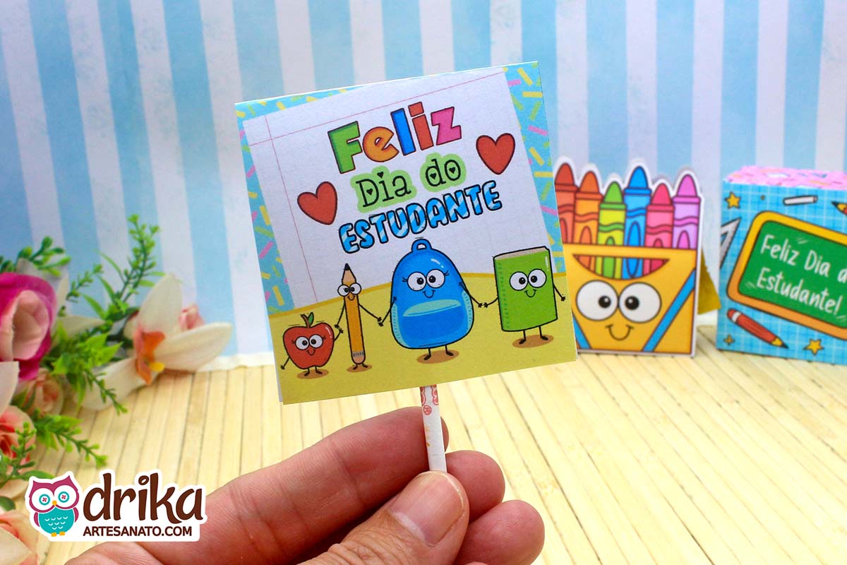 Mão segurando capinha de pirulito do Dia do Estudante com desenho de maçã, lápis, mochila e livro em fundo azul listrado