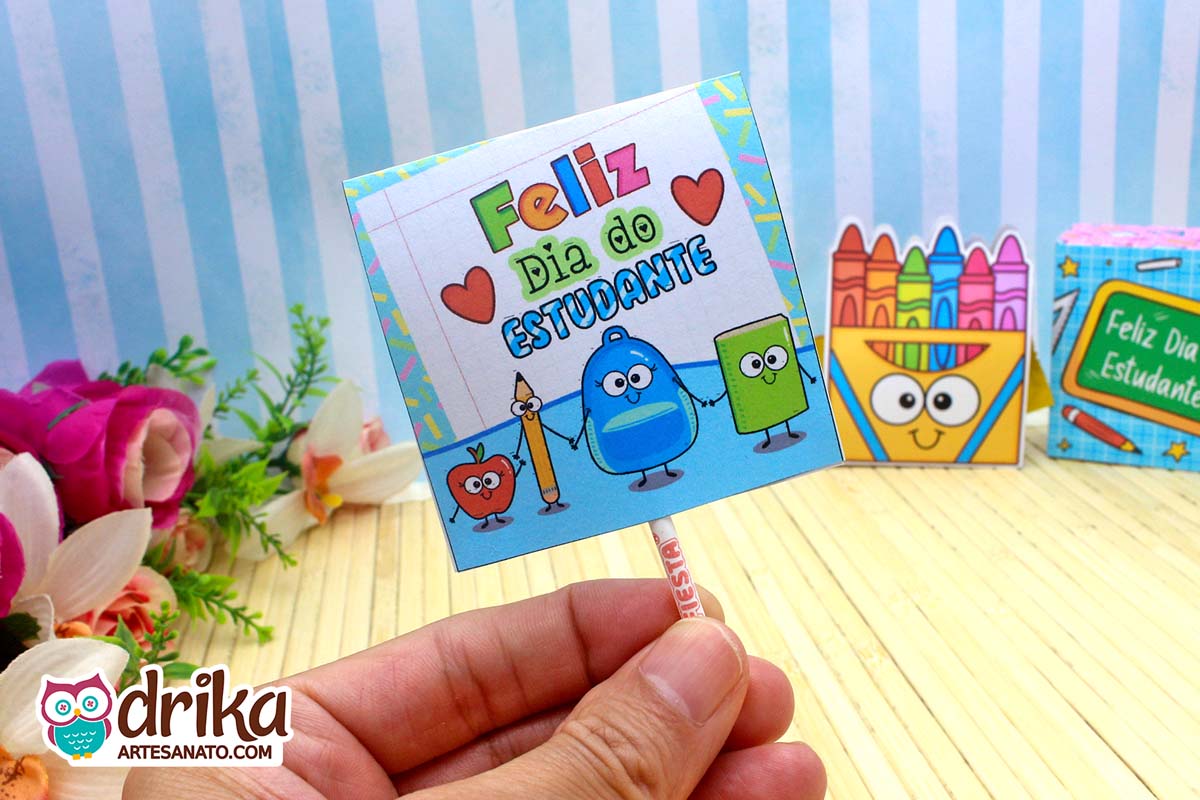 Detalhe de mão segurando capinha de pirulito do Dia do Estudante com personagens escolares em fundo azul listrado