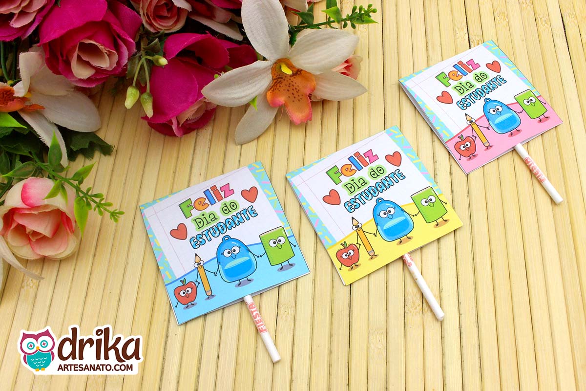 Três capinhas de pirulito do Dia do Estudante com personagens escolares sobre mesa de bambu e flores ao fundo