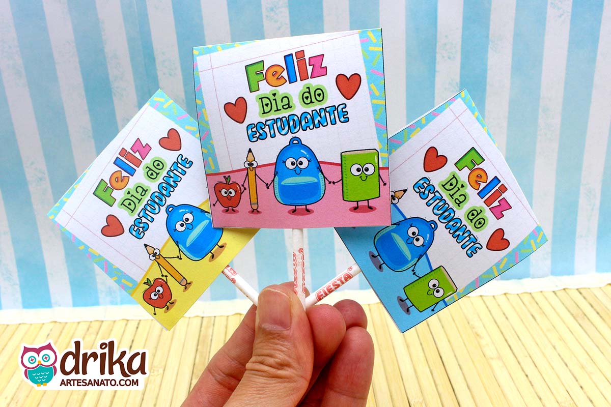 Mão segurando três capinhas de pirulito do Dia do Estudante com personagens escolares em fundo azul listrado