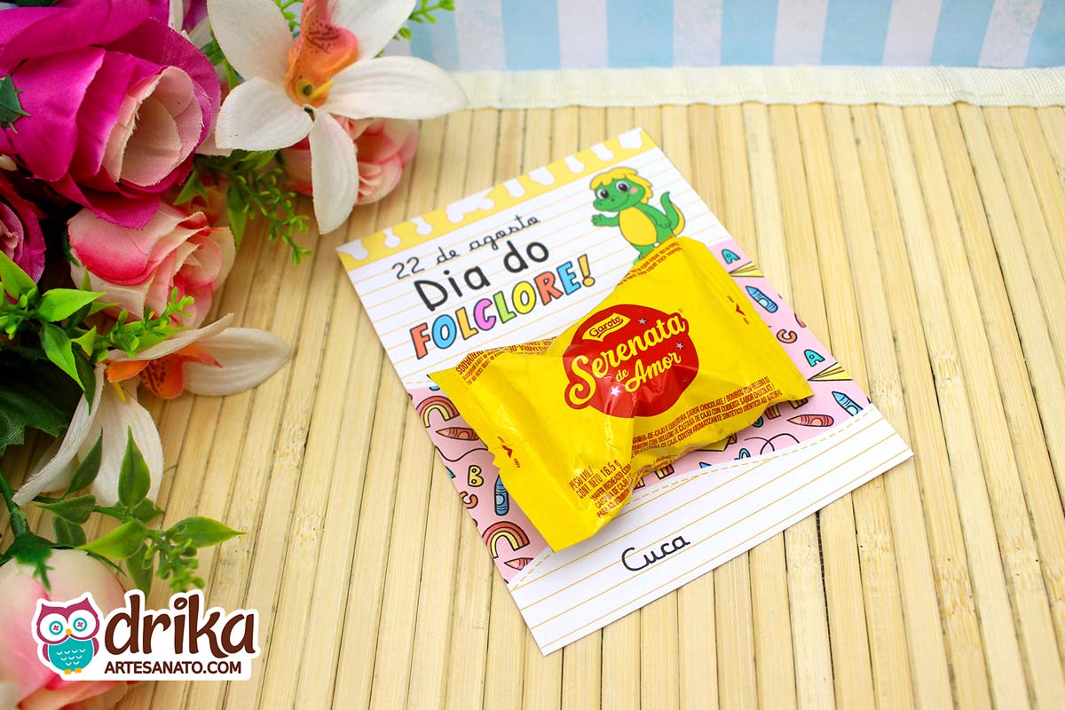 Cartão porta-bombom da Cuca com bombom Serenata de Amor, sobre mesa de bambu e flores.