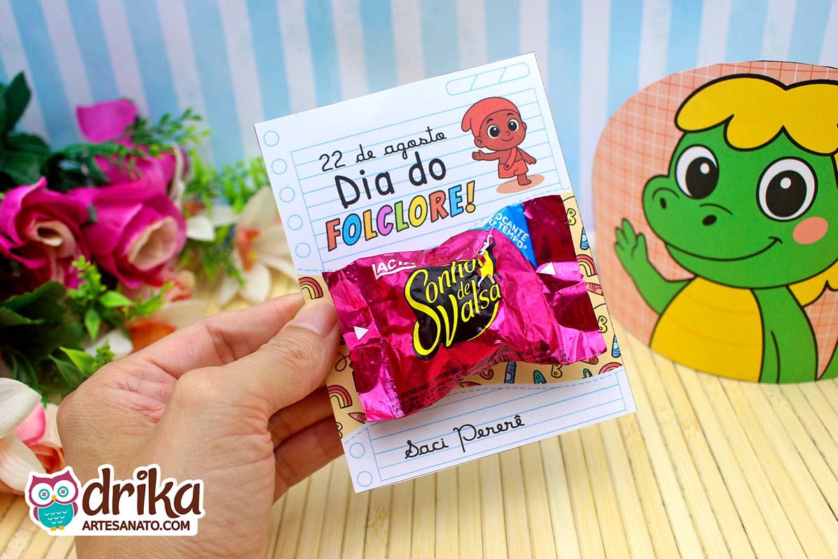 Mão segurando cartão porta-bombom do Saci Pererê com Sonho de Valsa, fundo com Cuca e flores.