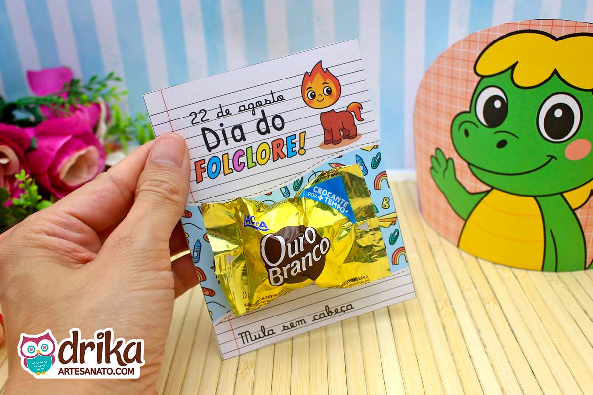 Mão segura cartão da Mula sem Cabeça com Ouro Branco, fundo com Cuca e flores.