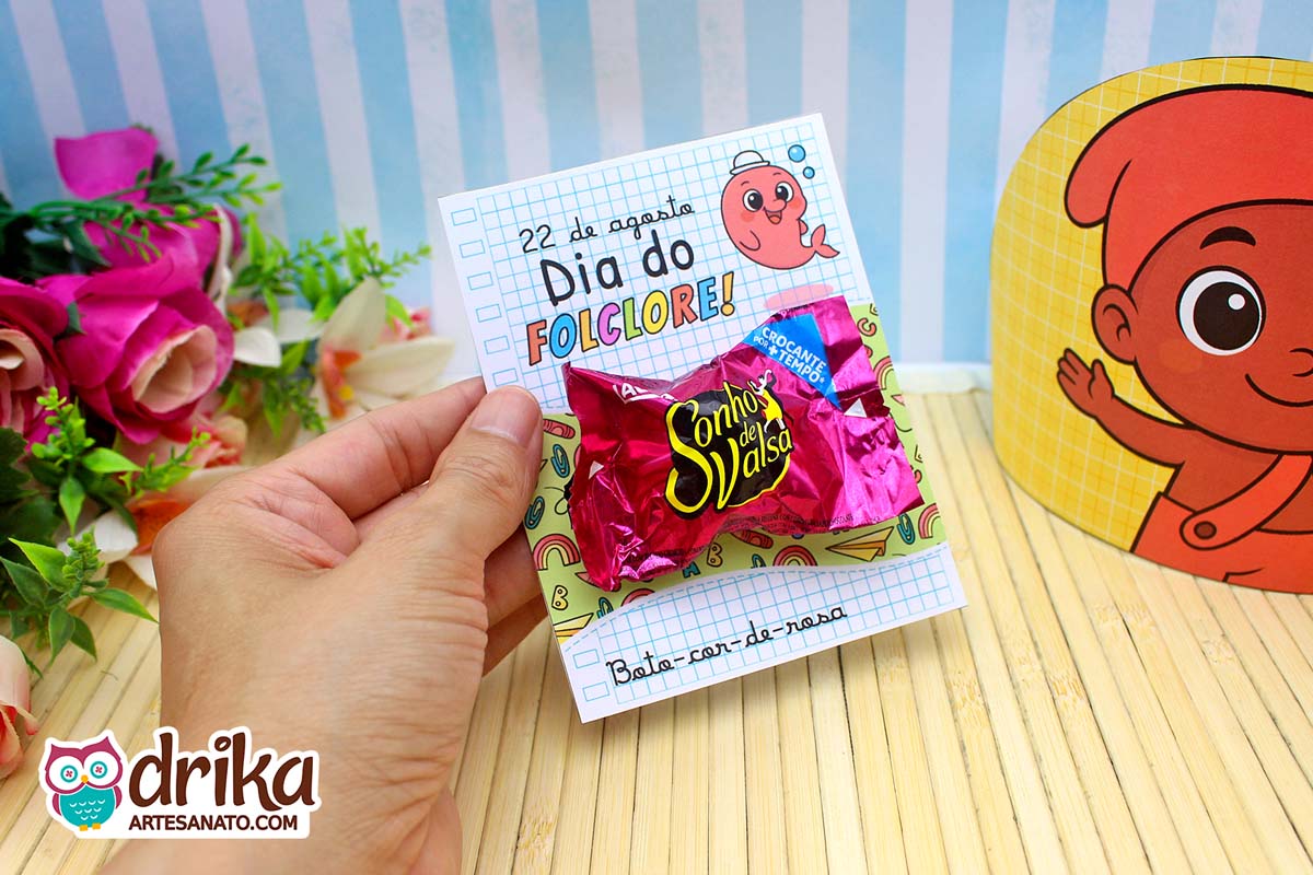 Mão segurando cartão do Boto-cor-de-rosa com Sonho de Valsa, fundo com Saci e flores.