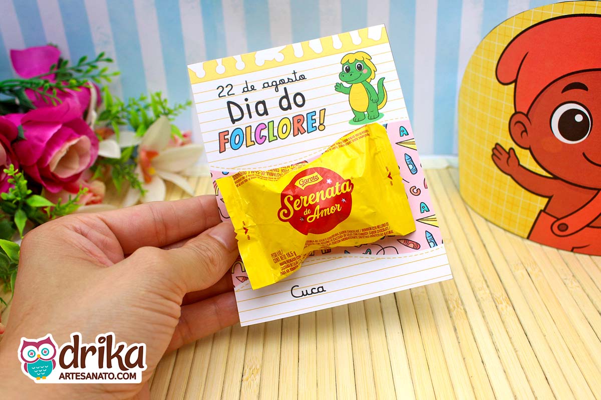 Mão segurando cartão da Cuca com Serenata de Amor, fundo com Saci e flores.