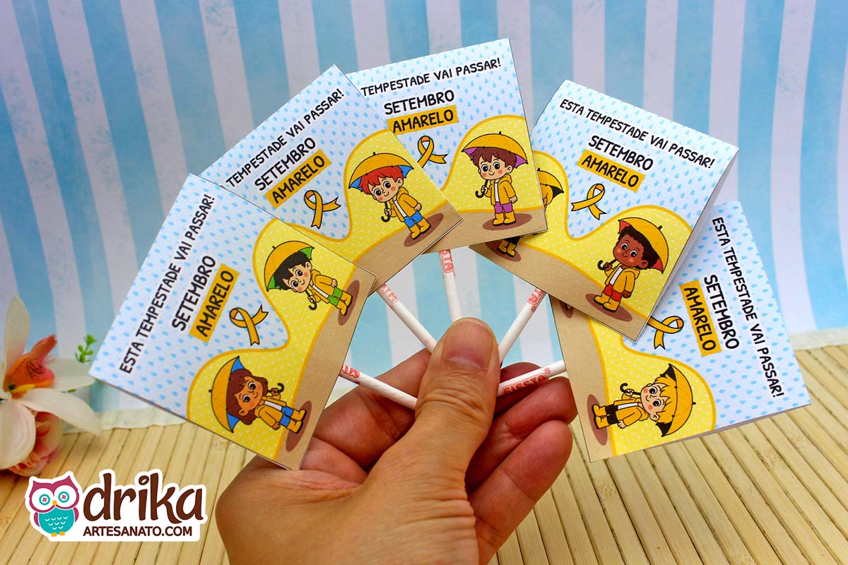 Mão segurando cinco capinhas de pirulito do Setembro Amarelo com desenhos de crianças protegidas da chuva.