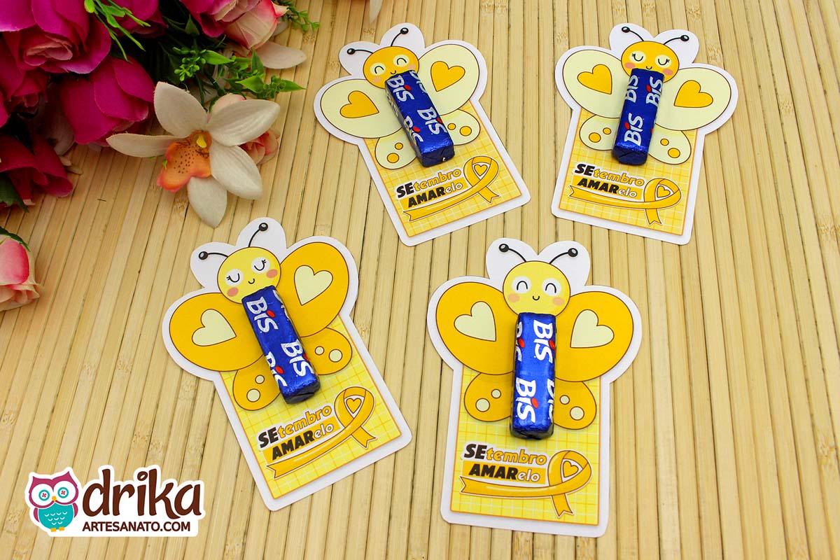 Quatro cartões porta-bis de borboleta amarelos com chocolate Bis azul em mesa de bambu e flores ao fundo