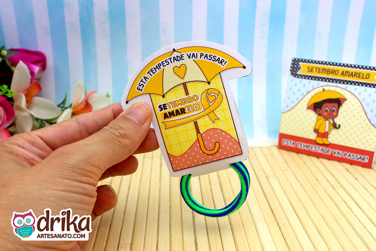 Porta-xuxinha guarda-chuva amarelo com frase motivacional, segurado por mão, com xuxinha verde e azul. Detalhes em vermelho.