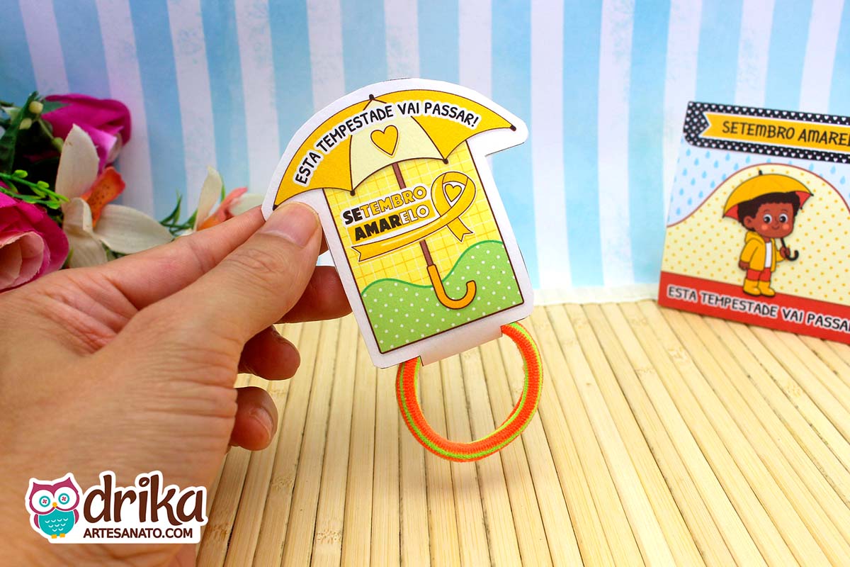 Mão segurando porta-xuxinha guarda-chuva verde do Setembro Amarelo com xuxinha laranja e verde. Frase "Esta tempestade vai passar".