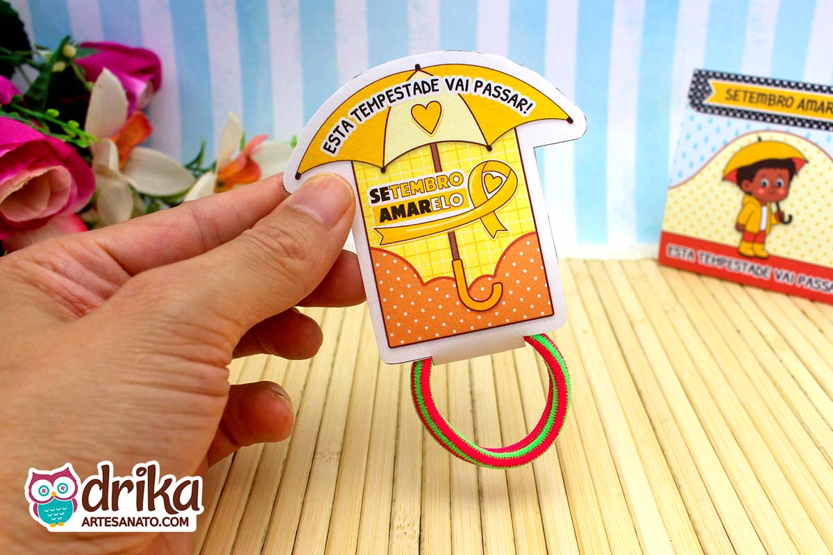 Mão segurando porta-xuxinha guarda-chuva laranja do Setembro Amarelo com xuxinha verde e rosa. Frase "Esta tempestade vai passar".