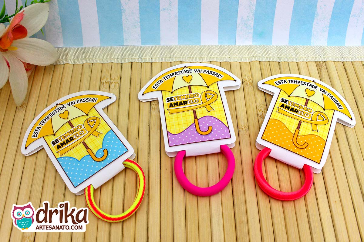 Três porta-xuxinhas guarda-chuva: azul, lilás e amarelo, com xuxinhas coloridas sobre mesa de bambu.