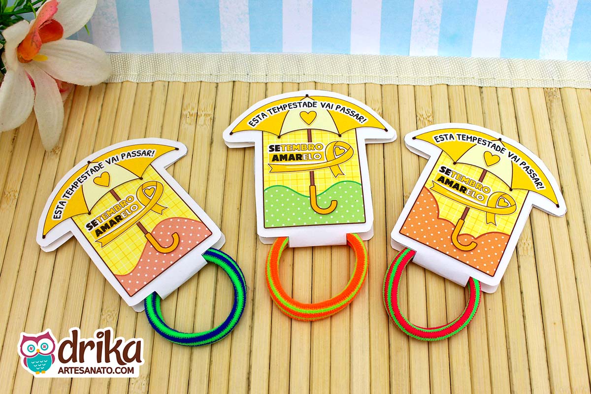 Três porta-xuxinhas guarda-chuva: vermelho, verde e laranja, com xuxinhas coloridas sobre mesa de bambu.