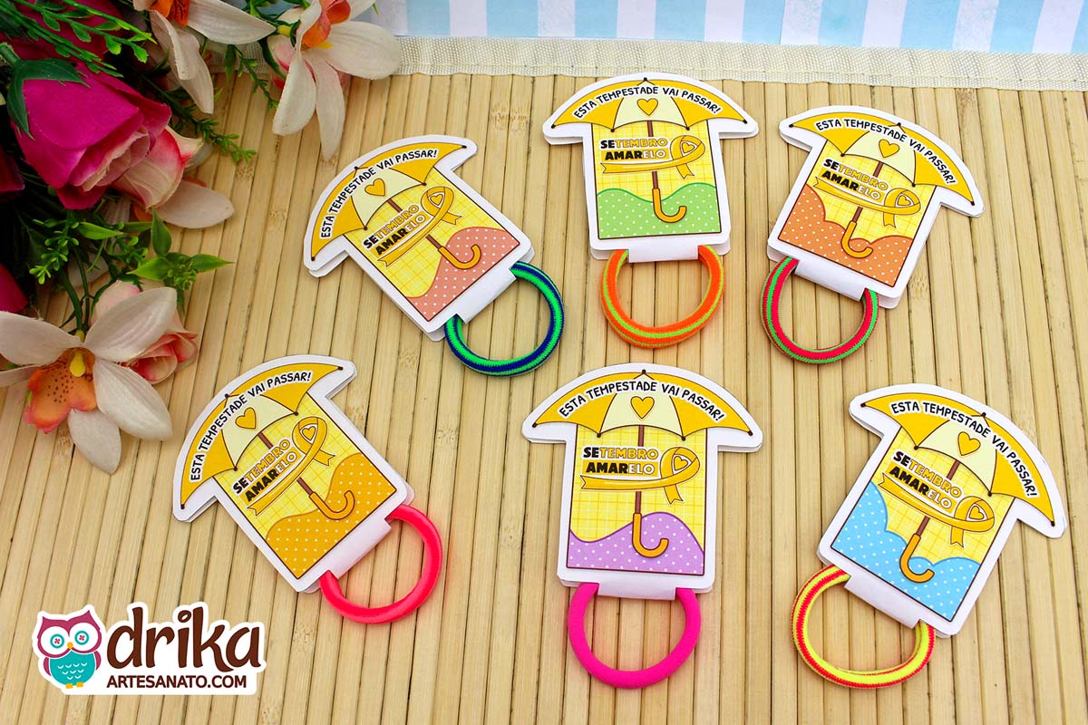 Seis porta-xuxinhas guarda-chuva do Setembro Amarelo com xuxinhas coloridas sobre mesa de madeira clara e flores ao fundo