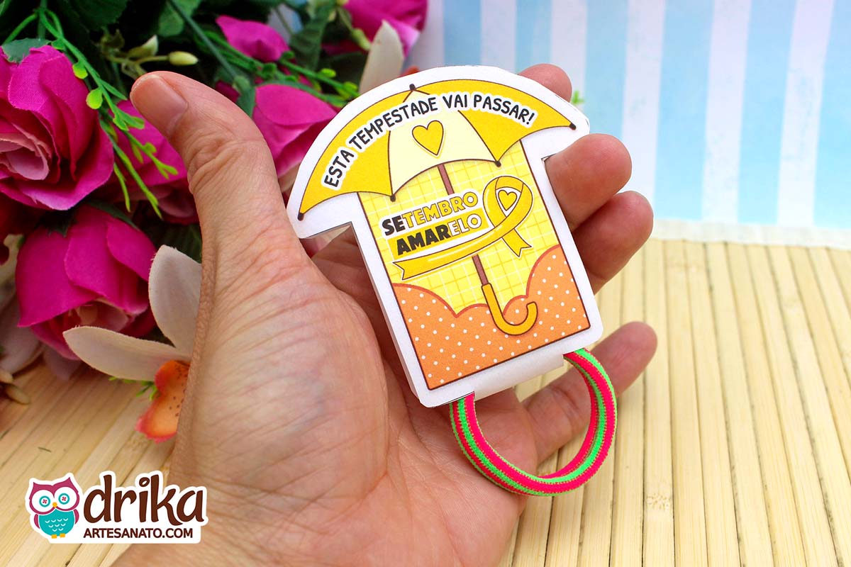 Porta-xuxinha guarda-chuva amarelo com detalhes laranja e frase motivacional, segurado por mão, com xuxinha verde e vermelha.