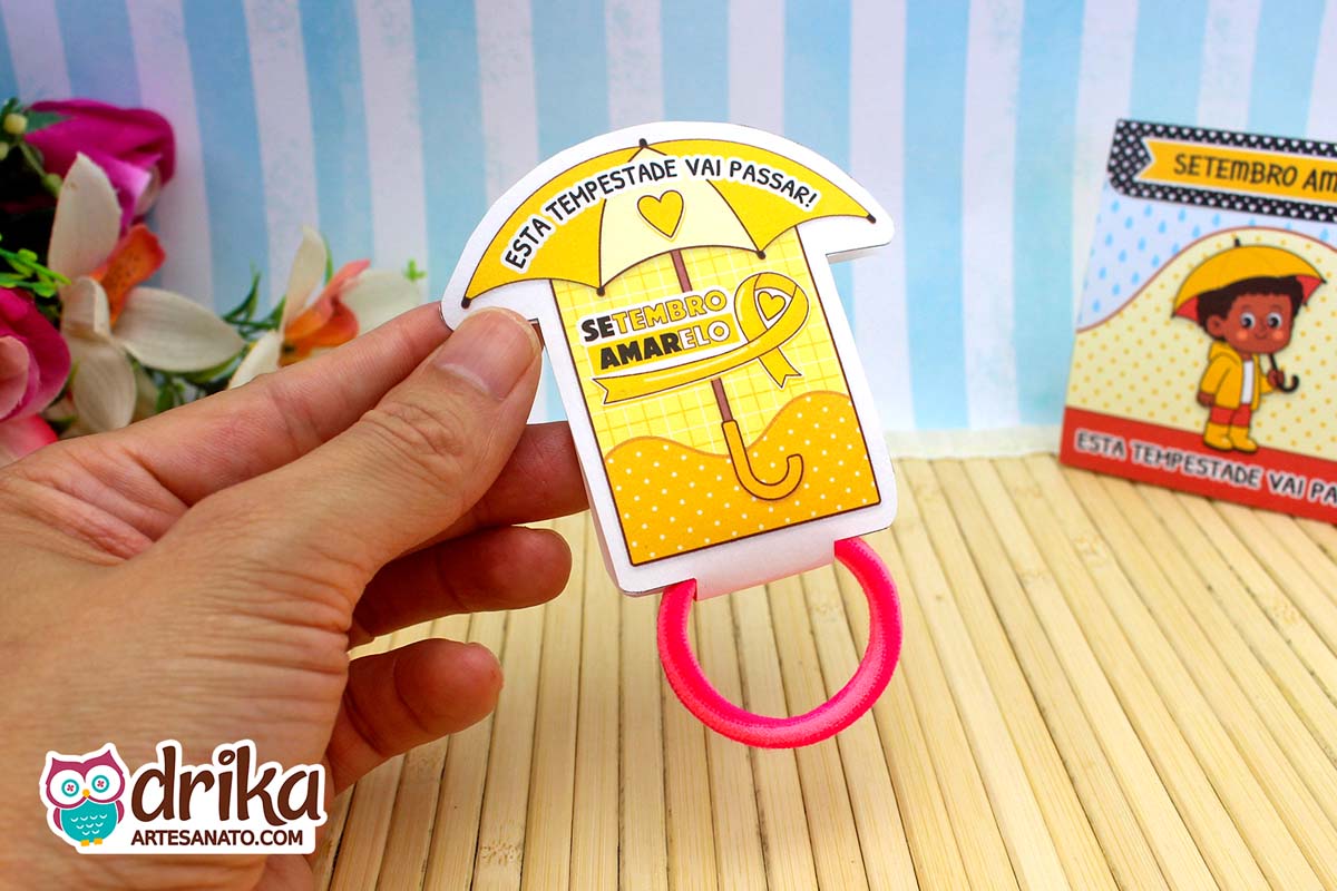 Porta-xuxinha guarda-chuva amarelo com frase motivacional, segurado por mão, com xuxinha rosa. Detalhes em amarelo.