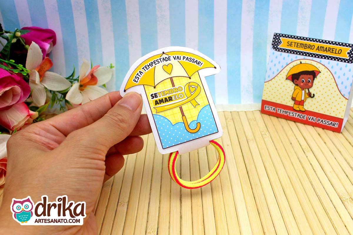 Porta-xuxinha guarda-chuva amarelo com frase motivacional, segurado por mão, com xuxinha amarela e vermelha. Detalhes em azul.