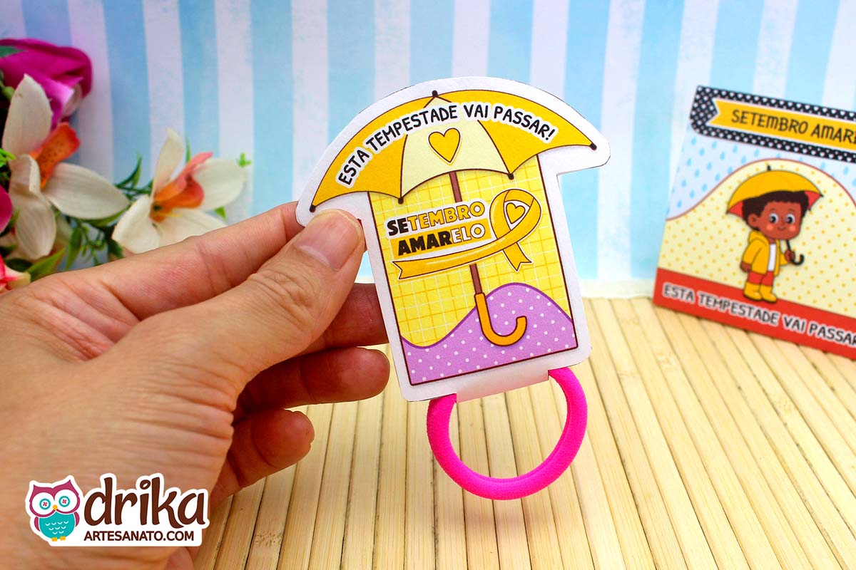 Porta-xuxinha guarda-chuva amarelo com frase motivacional, segurado por mão, com xuxinha rosa pink. Detalhes em lilás.