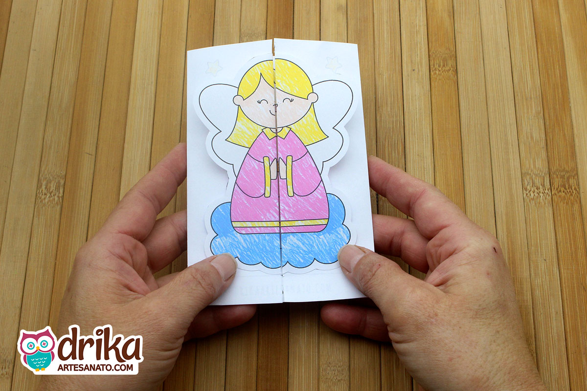 Mãos seguram cartão de Natal fechado com anjinha de vestido rosa e asas brancas sobre nuvem azul.