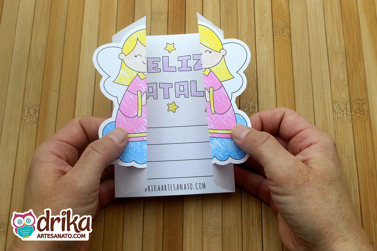 Mãos abrem cartão de Natal com anjinha, revelando mensagem "Feliz Natal" e espaço para escrever.