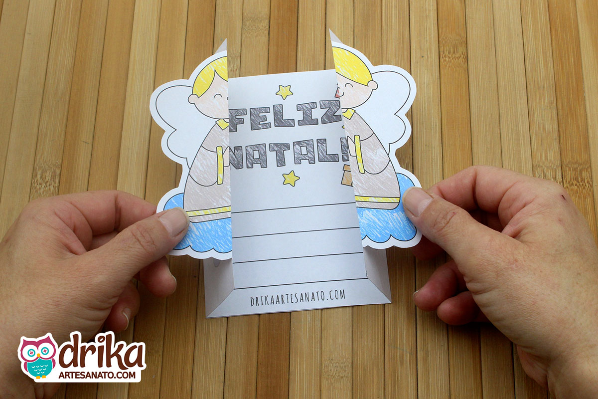 Mãos abrindo cartão de Natal com anjo, revelando mensagem "Feliz Natal" e espaço para escrever