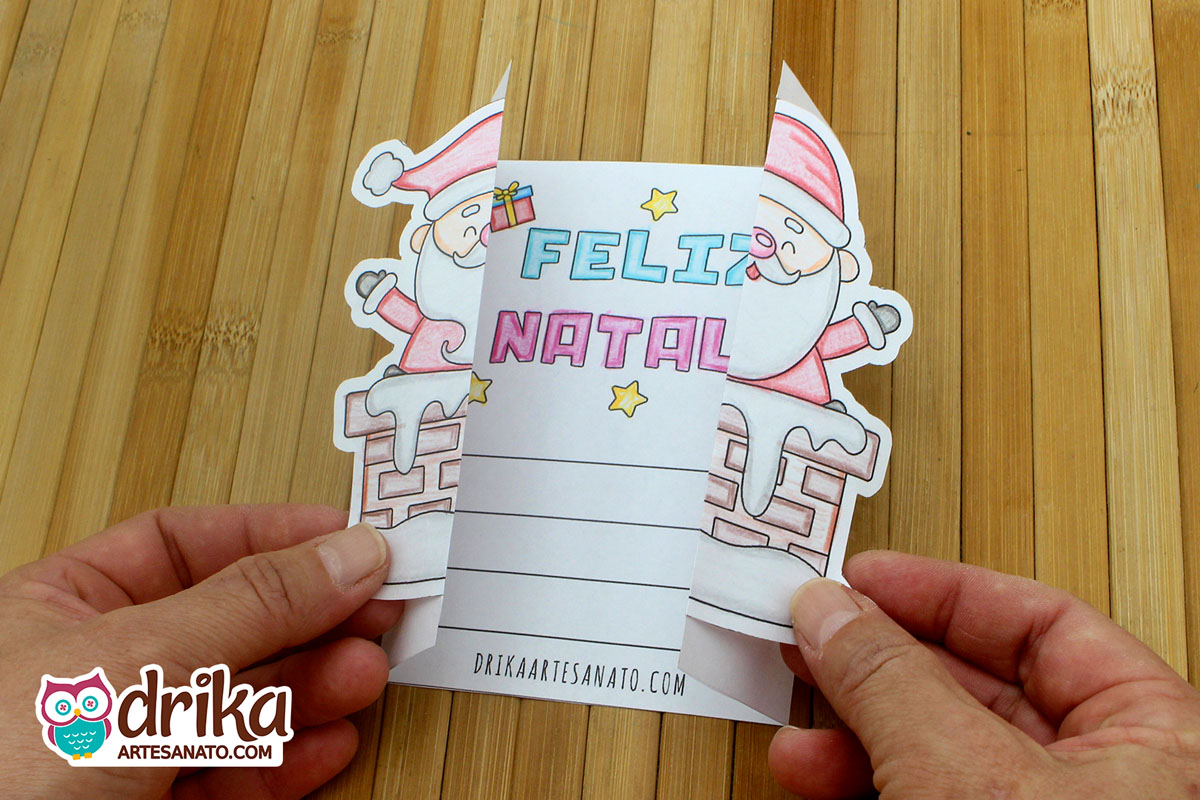 Mãos abrindo cartão de Natal com Papai Noel na chaminé, revelando mensagem de "Feliz Natal" e espaço para escrever