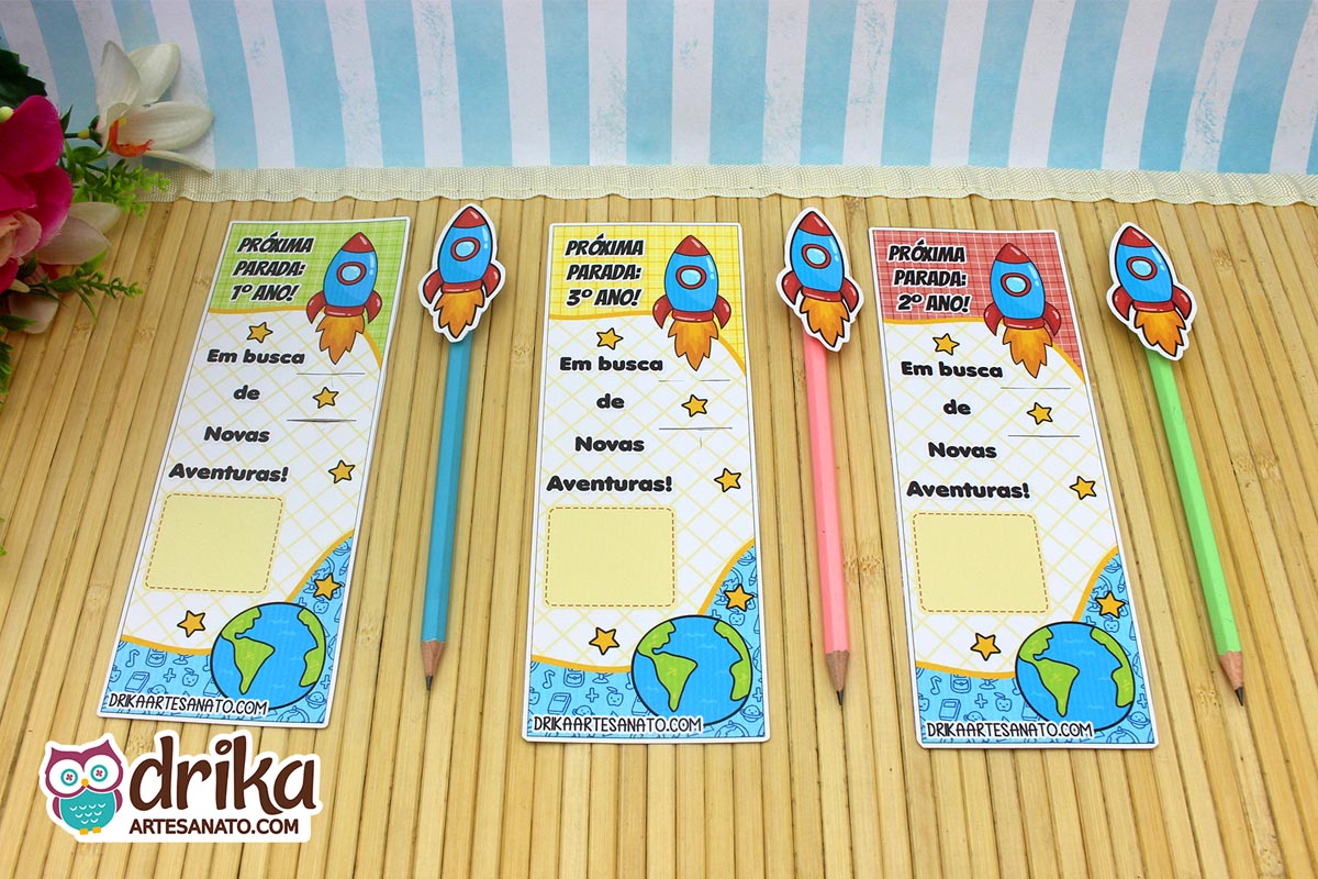 Três cartões porta-lápis de foguete para lembrancinha de férias, com lápis coloridos ao lado, em fundo de madeira clara.
