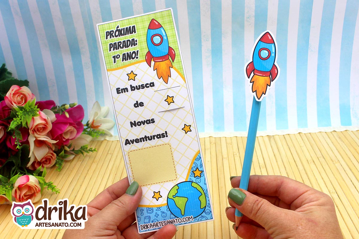 Cartão porta-lápis de foguete para o 1º ano, com lápis azul, sendo segurado por mãos com unhas verdes.