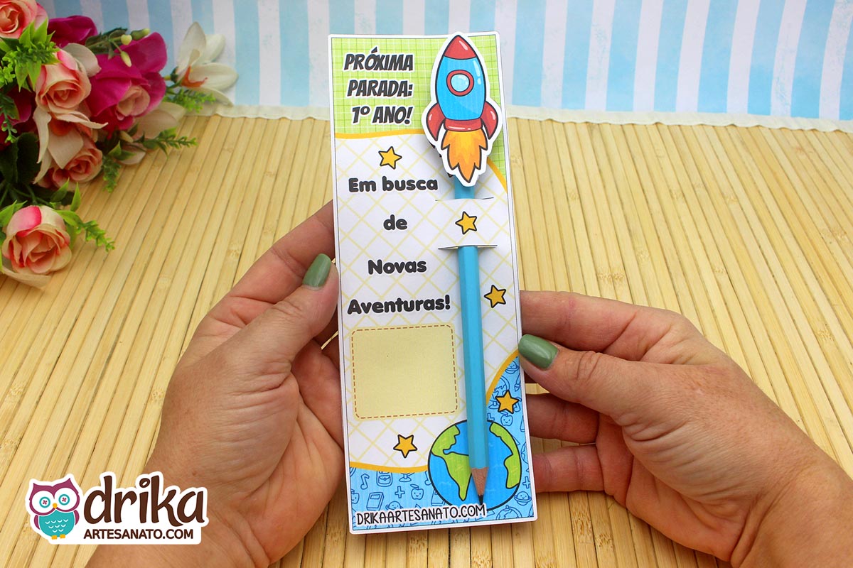 Mãos segurando cartão porta-lápis de foguete do 1º ano com lápis azul encaixado, simulando a propulsão.