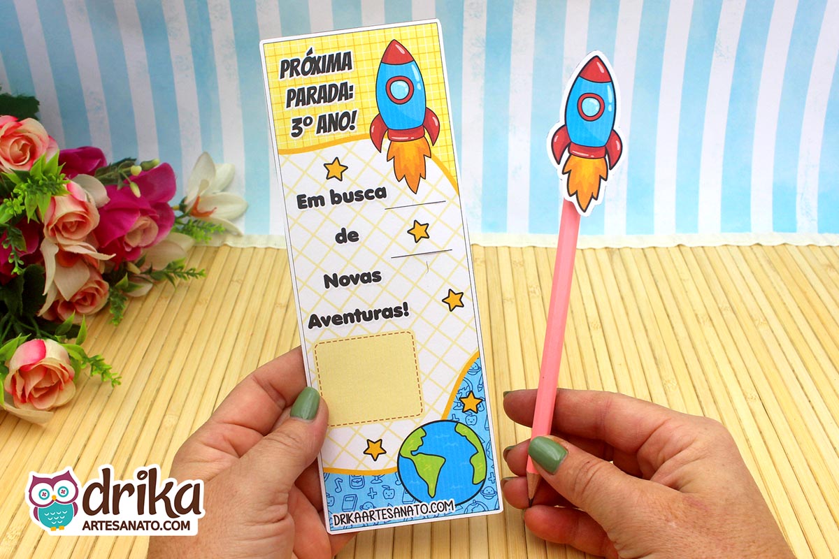 Mãos seguram cartão porta-lápis de foguete para o 3º ano, com foguete, estrelas e mensagem "Em busca de Novas Aventuras"