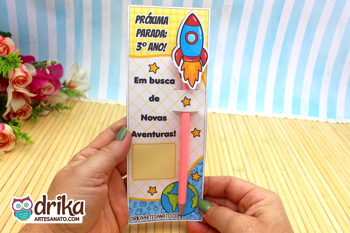 Mãos seguram cartão porta-lápis de foguete do 3º ano com lápis rosa encaixado, simulando a propulsão do foguete