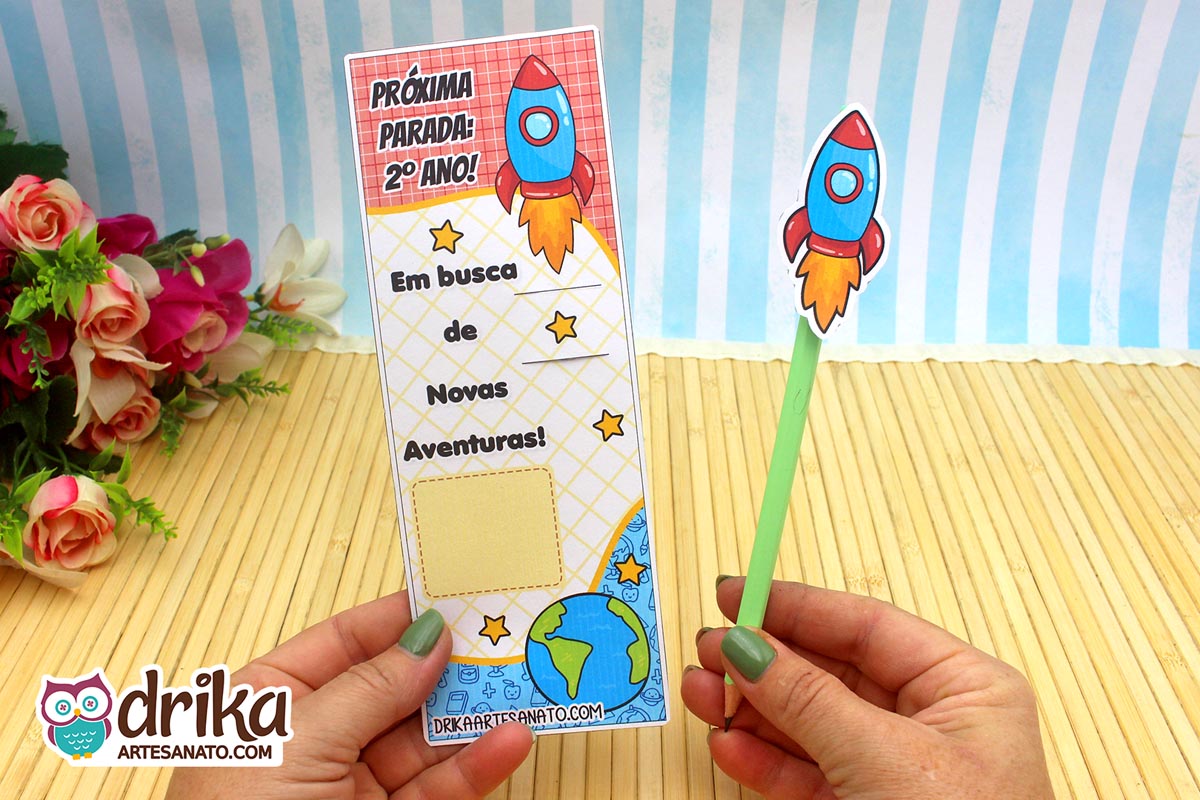 Cartão porta-lápis de foguete para o 2º ano, com lápis verde, sendo segurado por mãos com unhas verdes.