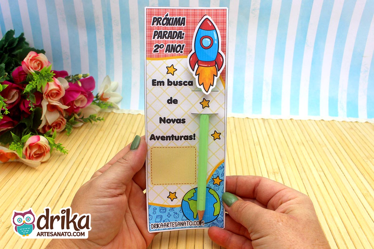 Mãos segurando cartão porta-lápis de foguete do 2º ano com lápis verde encaixado, simulando a decolagem.