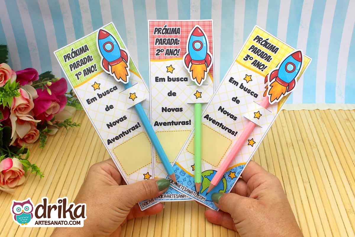 Mãos seguram três cartões porta-lápis de foguete para 1º, 2º e 3º ano, com lápis coloridos e design temático