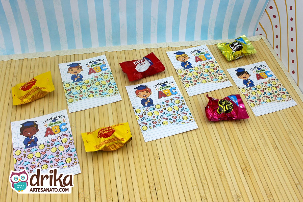 Cinco cartões porta-bombom de formatura ABC com desenhos de meninos e bombons variados sobre mesa de madeira.