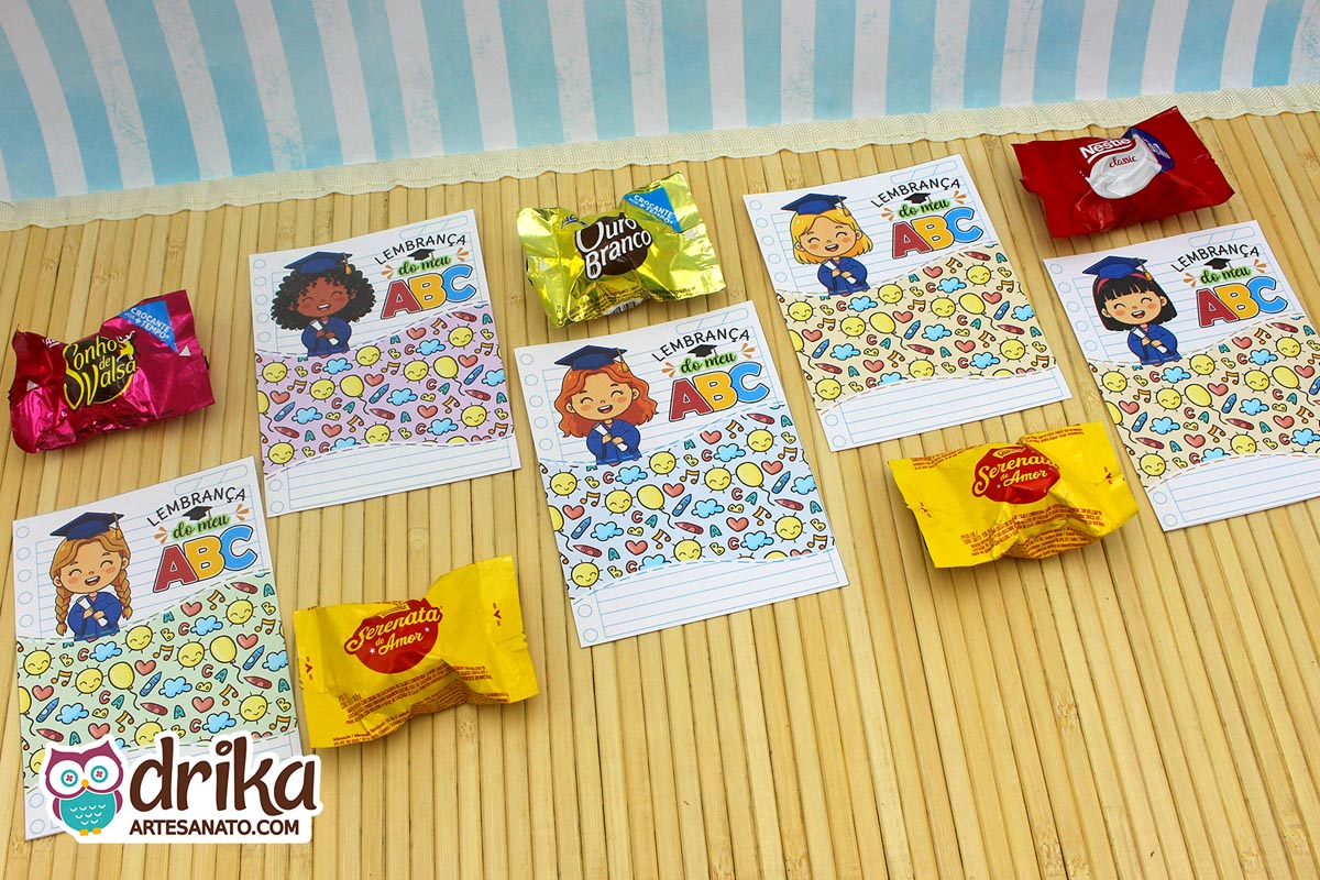 Cinco cartões porta-bombom de formatura ABC com meninas de diferentes etnias e bombons variados sobre mesa de bambu