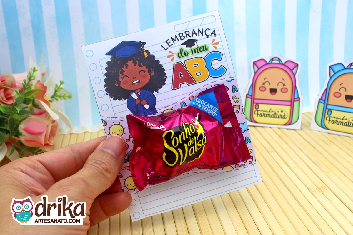 Mão segurando cartão porta-bombom Formatura ABC com menina de cabelo crespo e bombom Sonho de Valsa