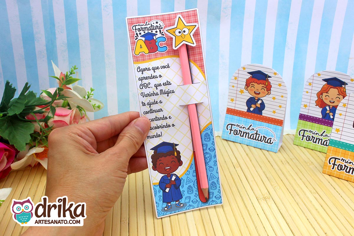 Mão segurando cartão porta-lápis formatura ABC com menino de cabelo crespo, lápis rosa com estrela e frase motivacional.
