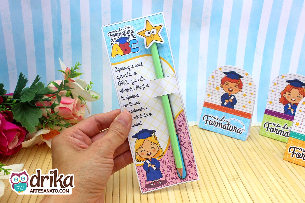 Mão segurando cartão porta-lápis Formatura ABC com menina loira e lápis/varinha verde com estrela amarela.