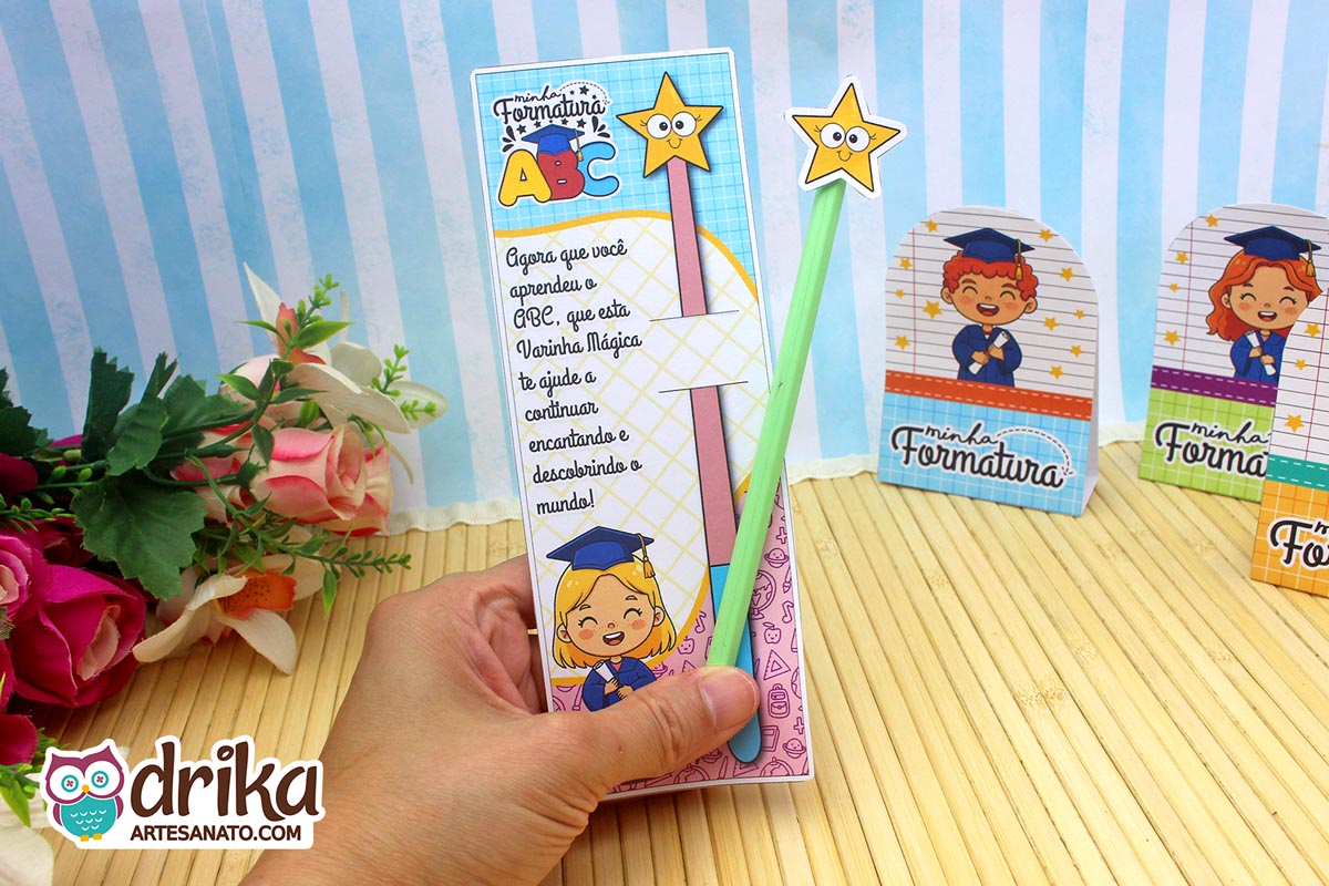 Mão segurando cartão porta-lápis formatura ABC com menina loira, lápis verde com estrela amarela, frase inspiradora e desenhos temáticos.