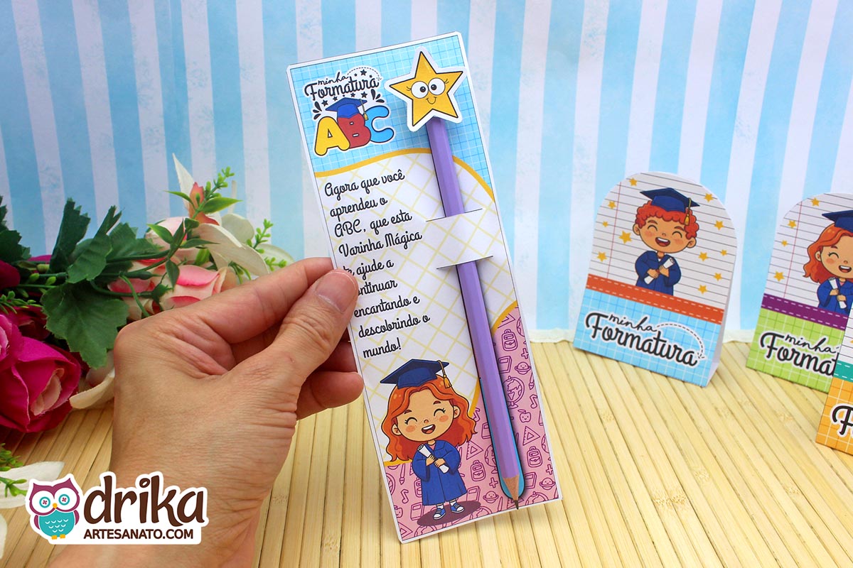 Mão segurando cartão formatura ABC com menina ruiva, lápis roxo com estrela, frase inspiradora e desenhos.