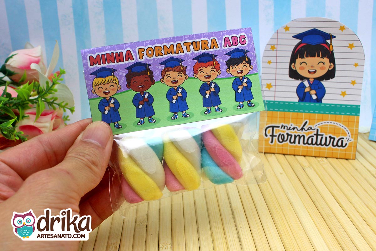 Mão segurando saquinho com marshmallows e lapela roxa e verde de formatura ABC com desenhos de formandos.
