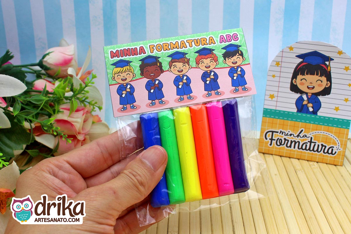 Mão segurando saquinho com massinhas e lapela verde e vermelha de formatura ABC com desenhos de formandos.