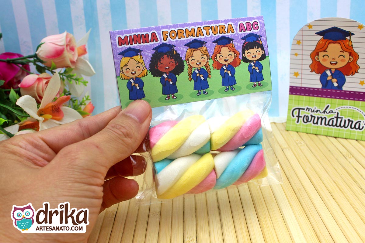 Mão segurando saquinho com marshmallow e lapela "Minha Formatura ABC" com meninas de beca azul, tons de roxo e verde.