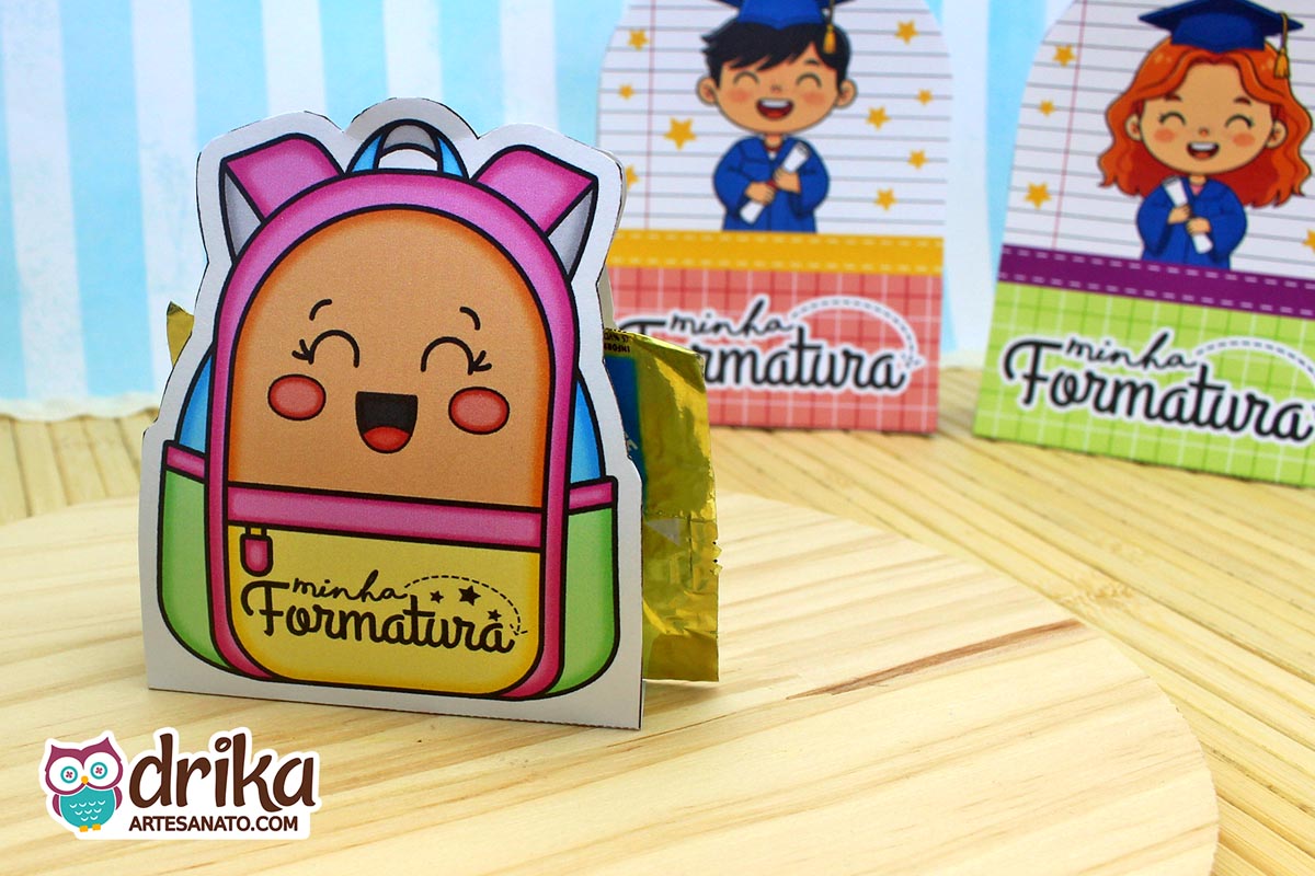 Mochila porta-bombom rosa e verde com rosto de menina sorrindo, bombom dourado à mostra e outras lembrancinhas ao fundo