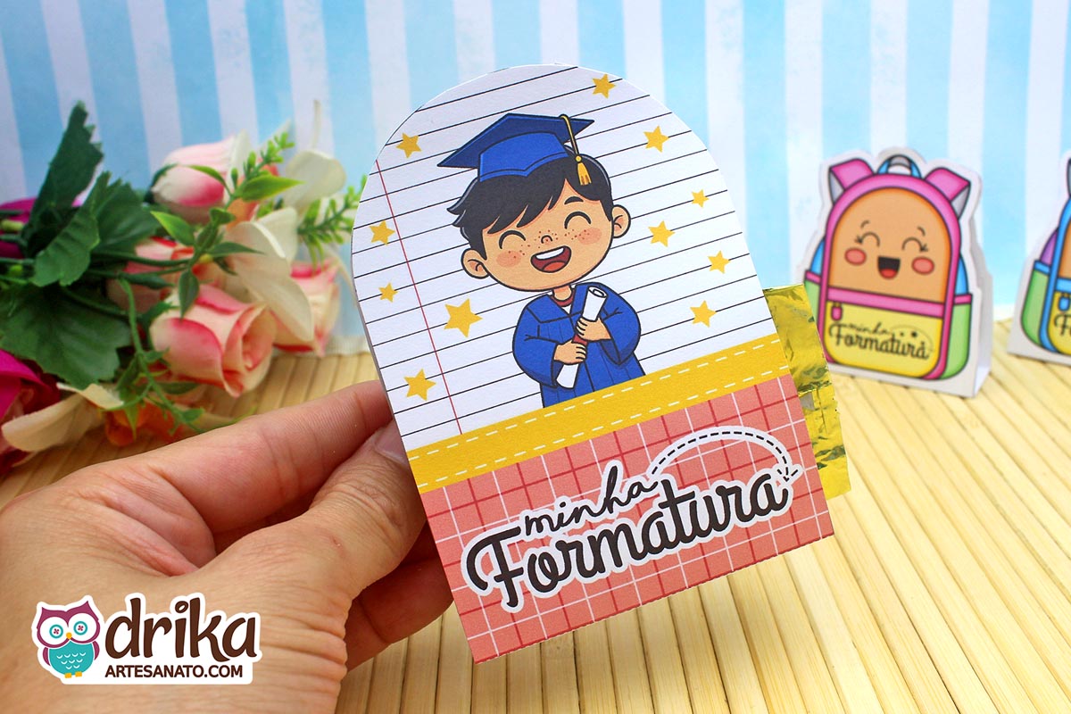 Mão segurando porta-bombom de formatura com menino de cabelo preto de beca azul e capelo, segurando diploma e fundo de caderno.