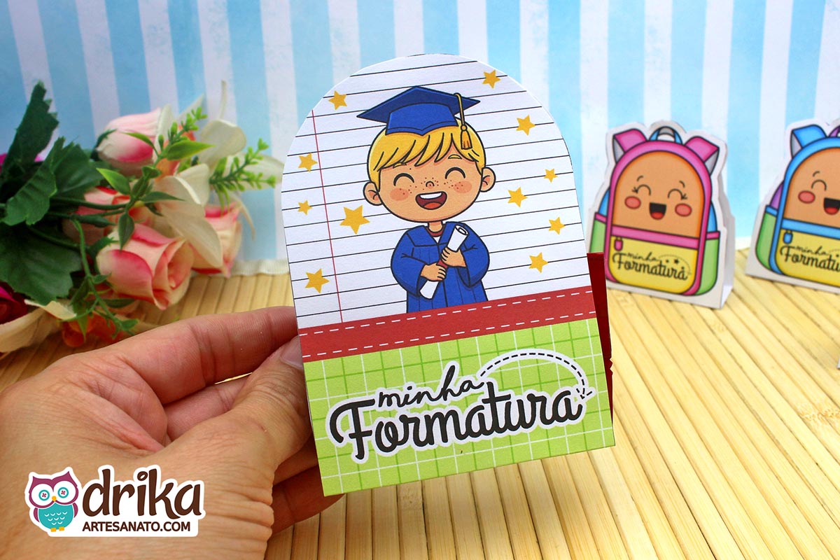 Mão segurando porta-bombom de formatura com menino loiro de beca azul e capelo, segurando diploma e fundo de caderno.