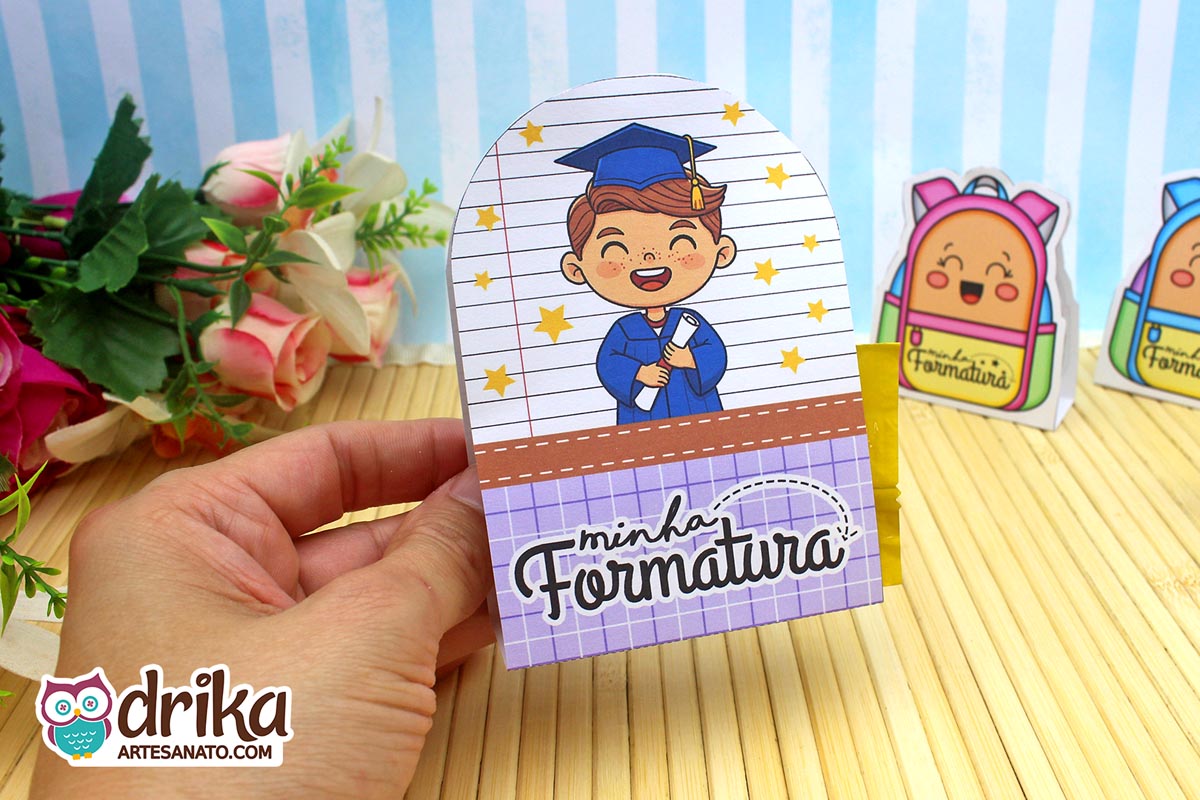 Mão segurando porta-bombom de formatura com menino castanho de beca azul e capelo, segurando diploma e fundo de caderno.