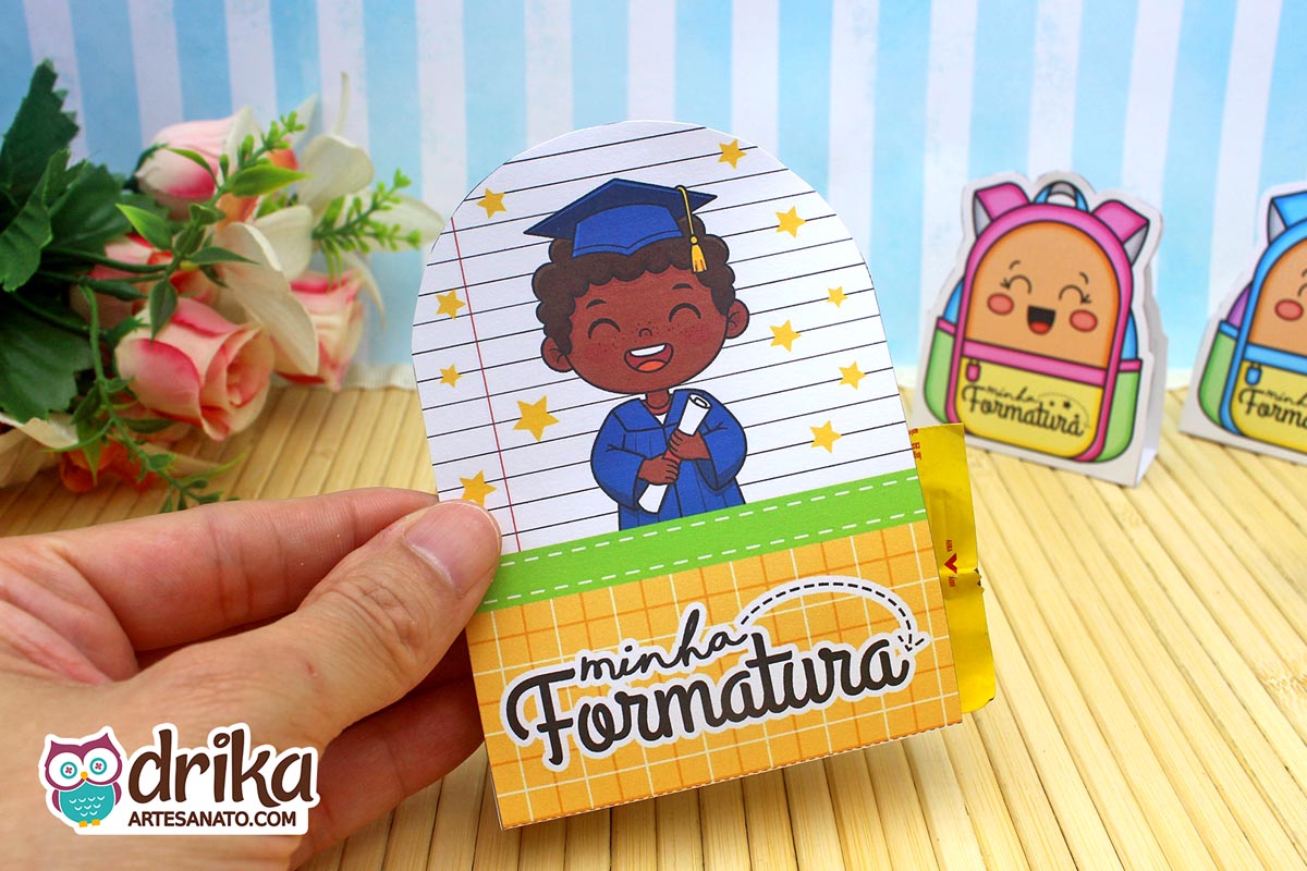 Mão segurando porta-bombom de formatura com menino negro de beca azul e capelo, segurando diploma e fundo de caderno.