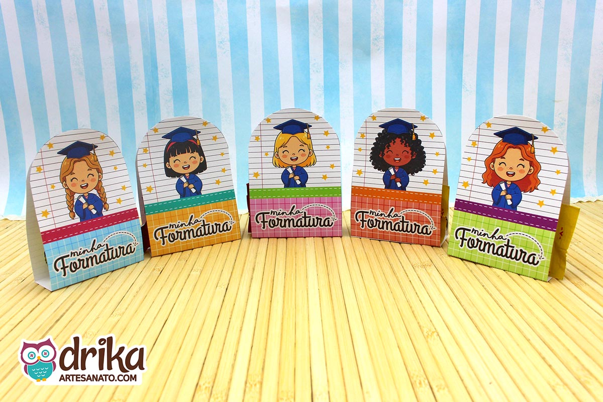 Cinco porta-bombons de formatura ABC com design de meninas cute em beca azul, lado a lado sobre mesa de bambu.