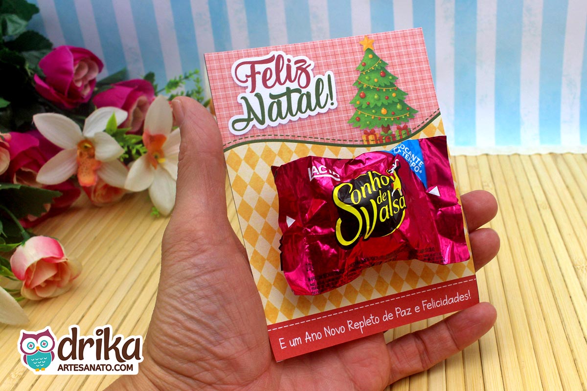 Mão segurando cartão porta-bombom de Natal com bombom Sonho de Valsa e fundo xadrez rosa.